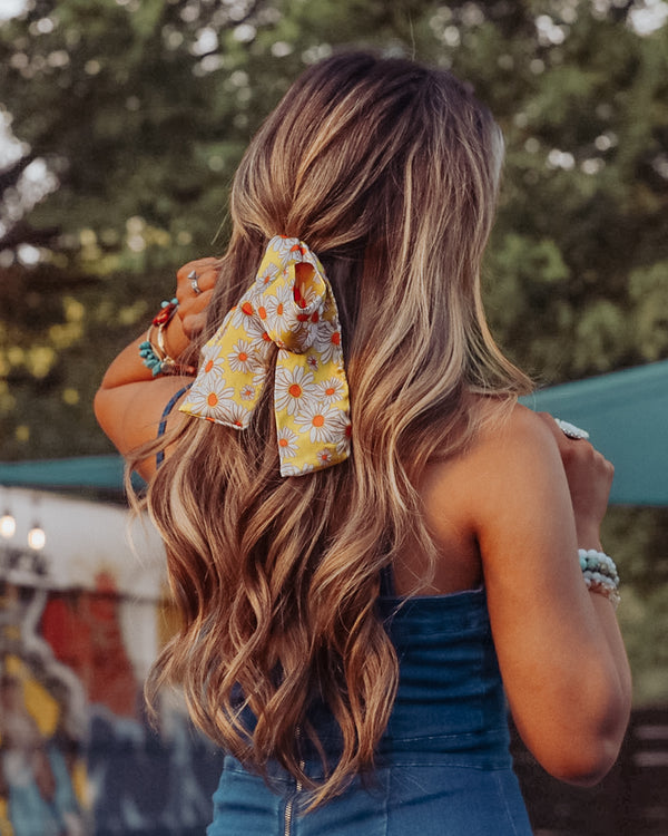 Image of brunette model with her hair styled by Beachwaver hair tool pulled away from face using a hair scarf that is yellow with white daisies