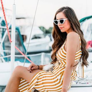 Image of model with long brunette hair styled by a Beachwaver pro 1 sitting on a doc with a yellow striped dress and white sunglasses.