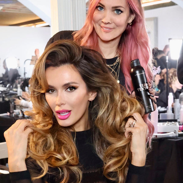 Image of Brunette Haired model that was styled into a glam voluminous look by Sarah Potempa CEO and Celebrity Hairstylist, holding up the team texture spray she used in the models hair  hair.